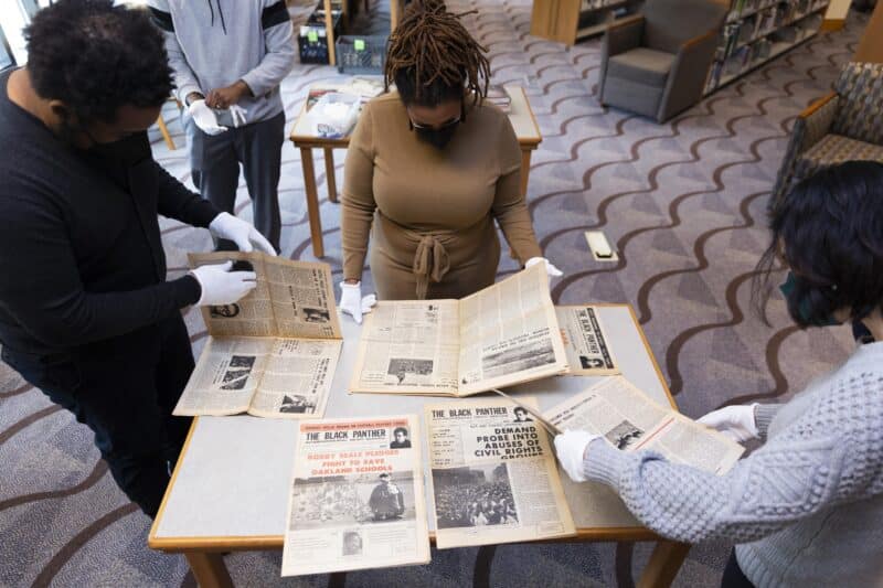 Three people wearing white gloves examine archival copies of newspapers from the Black Panther Party for Self-Defense.